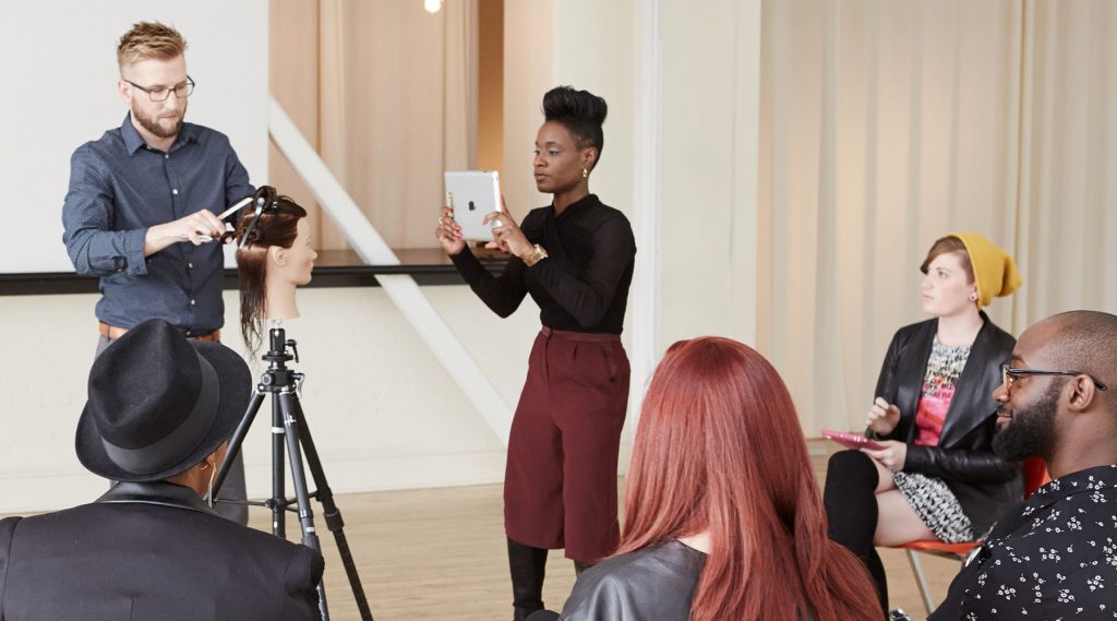 Pivot Point instructor showing students hairstyling techniques while another instructor recording with an ipad